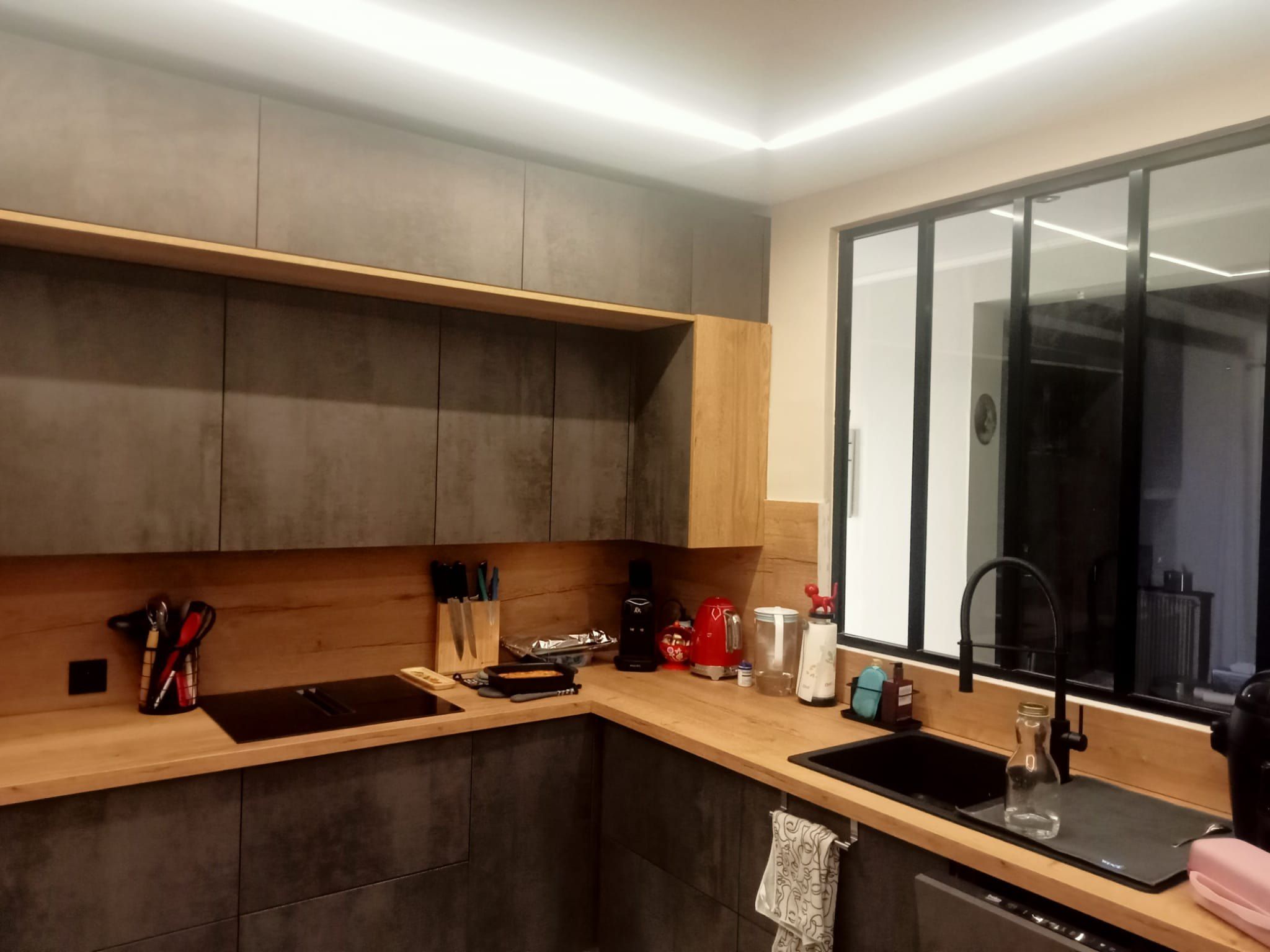La cuisine après travaux : façades effet béton, plan de travail et crédence en bois, plaque à induction, évier sous la verrière et corniche lumineuse
