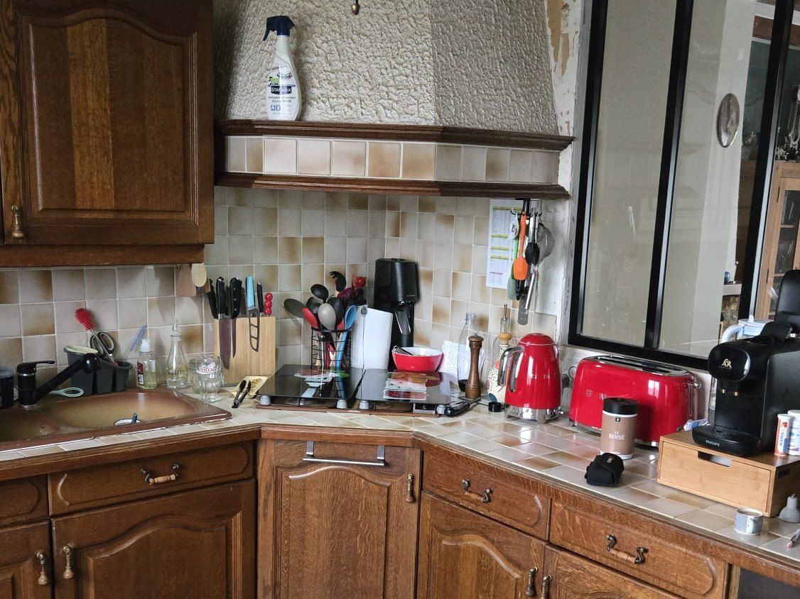 La même cuisine avant travaux : meubles en chêne massif, plan de travail carrelé, crédence en faïence beige et hotte maçonnée