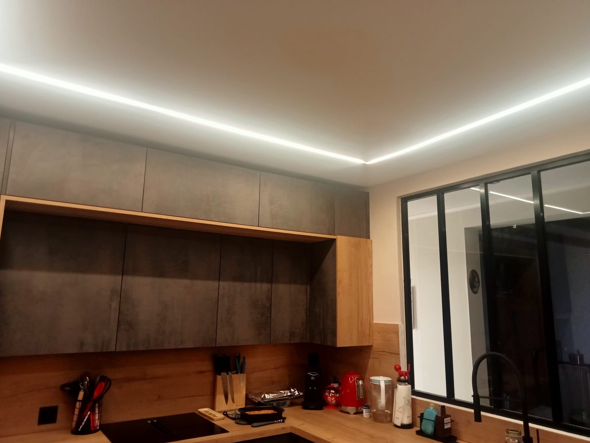 Cuisine en L après travaux : meubles hauts effet béton, corniche lumineuse courant sur tout le plafond et verrière atelier
