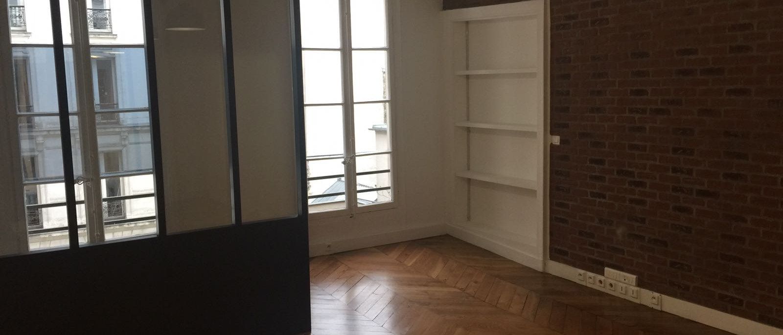 Séjour d'appartement rénové : parquet chevron, verrière atelier et mur de brique conservé