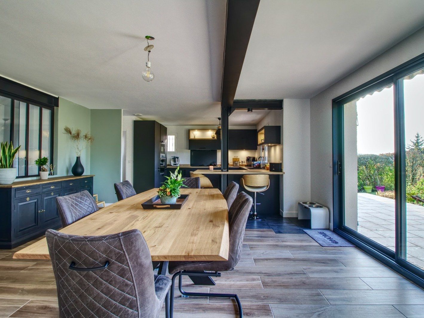 Salle à manger avec table bois massif, verrière et cuisine bleue en arrière-plan